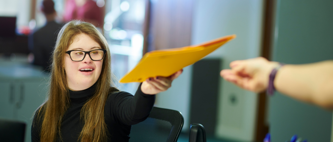 Woman with Down syndrome working in office handing a yellow file folder to coworker