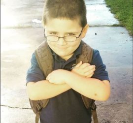 Elementary age boy with glasses wearing a backpack, dark green shirt, and folding his arm