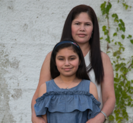 Hispanic mother and daughter standing in front of a white wall with green vines growing up it