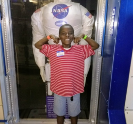 African American boy wearing red shirt and blue jean shorts standing in front of a NASA astronaut suit
