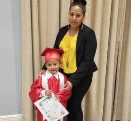 Hispanic mother wearing yellow shirt and black pantsuit with her kindergarten son wearing a red cap and gown and holding an award