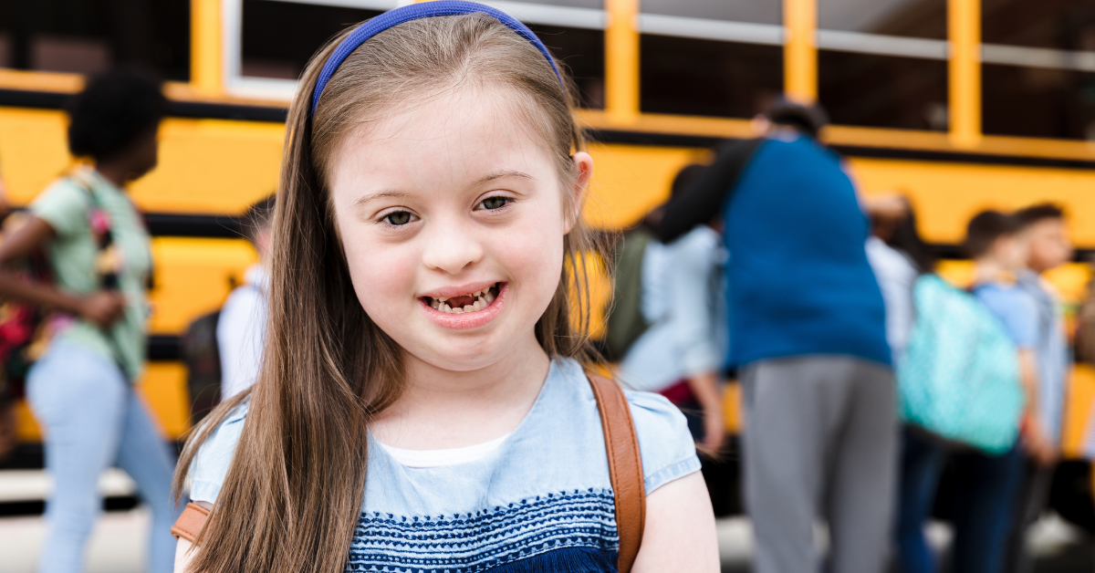 Elementary aged girl with down syndrome wearing a blue headband and blue dress standing in form of a school bus