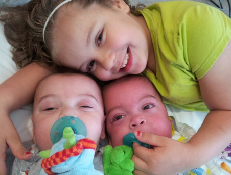 Young your with blonde hair and green shirt holding her twin infant brothers