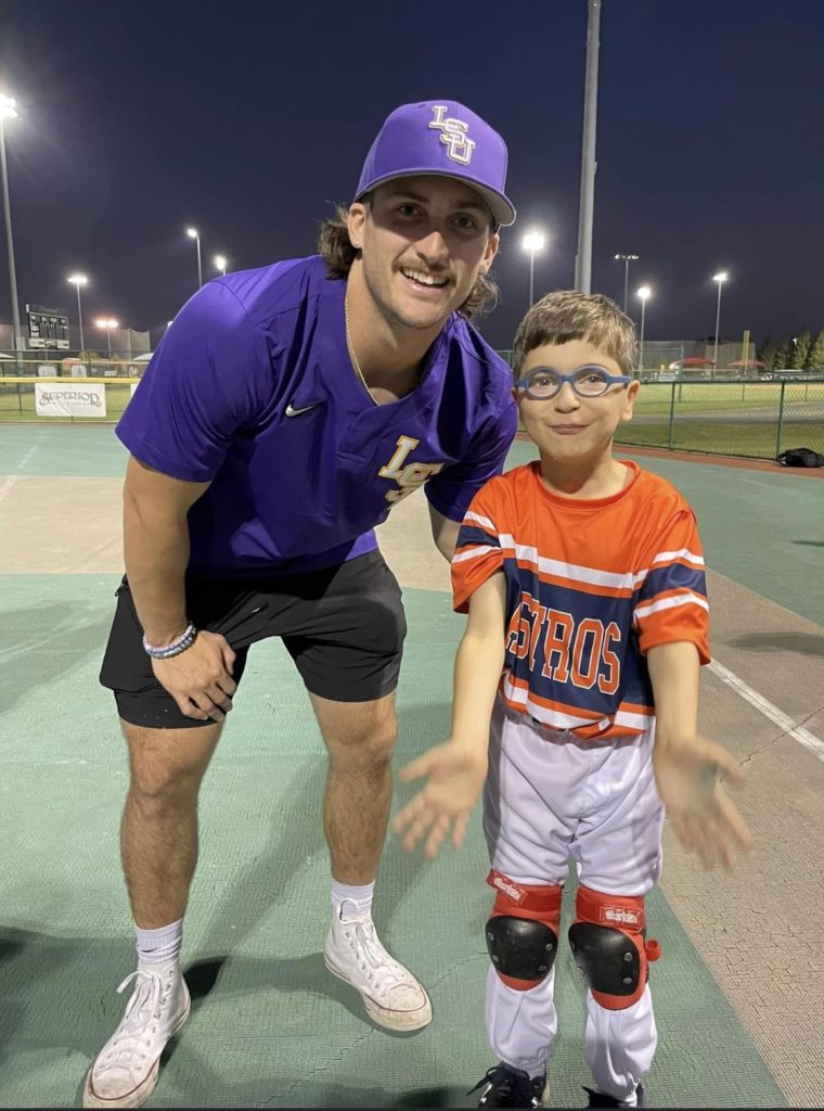 Dylan Crews wearing a baseball cap and LSU Shirt with young boy with a disability wearing glasses and orange white and blue shirt and white pants
