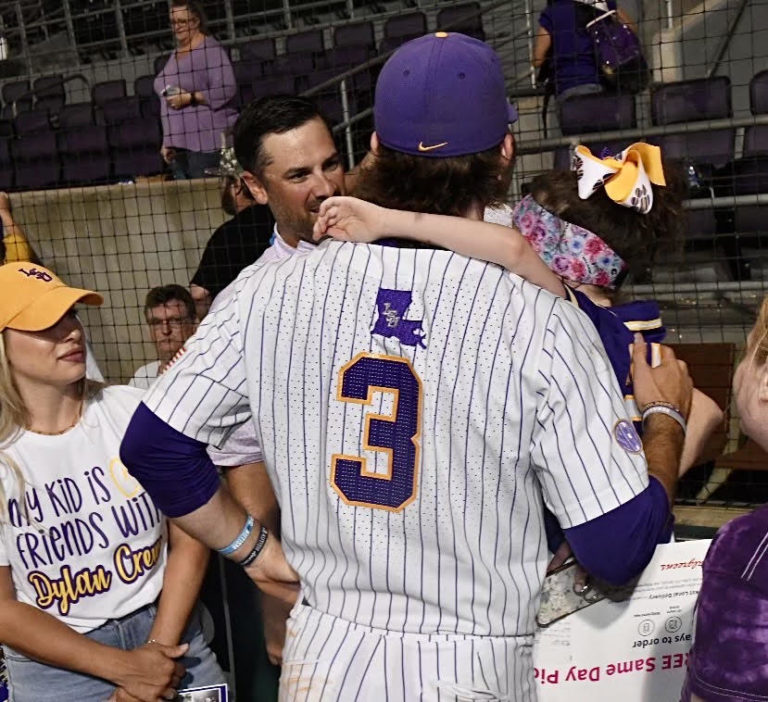 Woman with blond hair, yellow baseball cap, and shirt that say My kid is friends with Dylan Crews smiling . Back of Dylan Crews #3 LSU Baseball Player holding young child with a disability