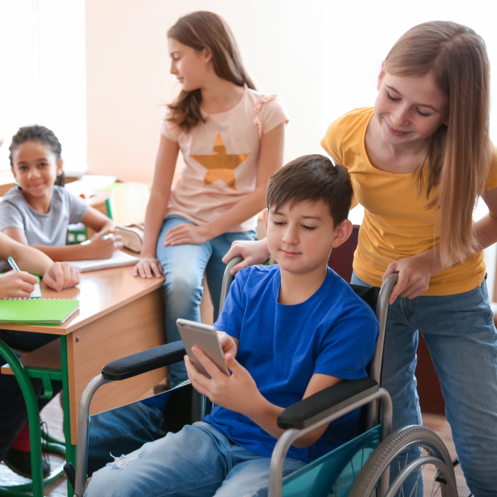 Middle school aged students in a classroom, girl looking over shoulder of boy in wheelchair at his device