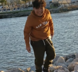 Hispanic male wearing orange sweatshirt and khaki pants standing on rocks by the lake