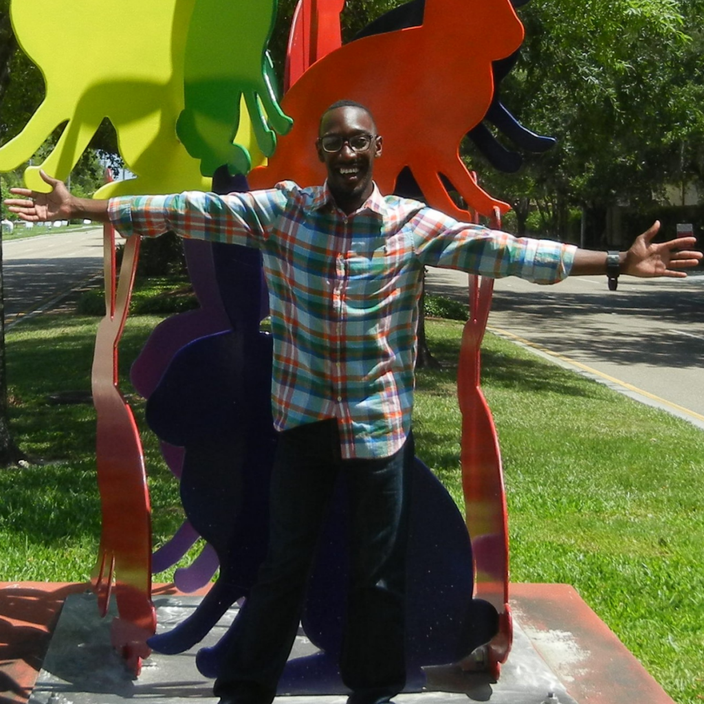 Man who is African American standing in front of art sculpture holding his arms wide open