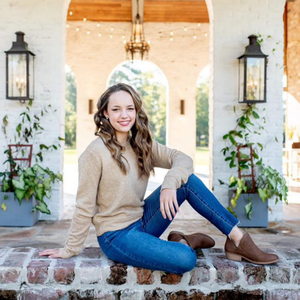 High School Girl wearing jeans and a tan sweater sitting on bricks